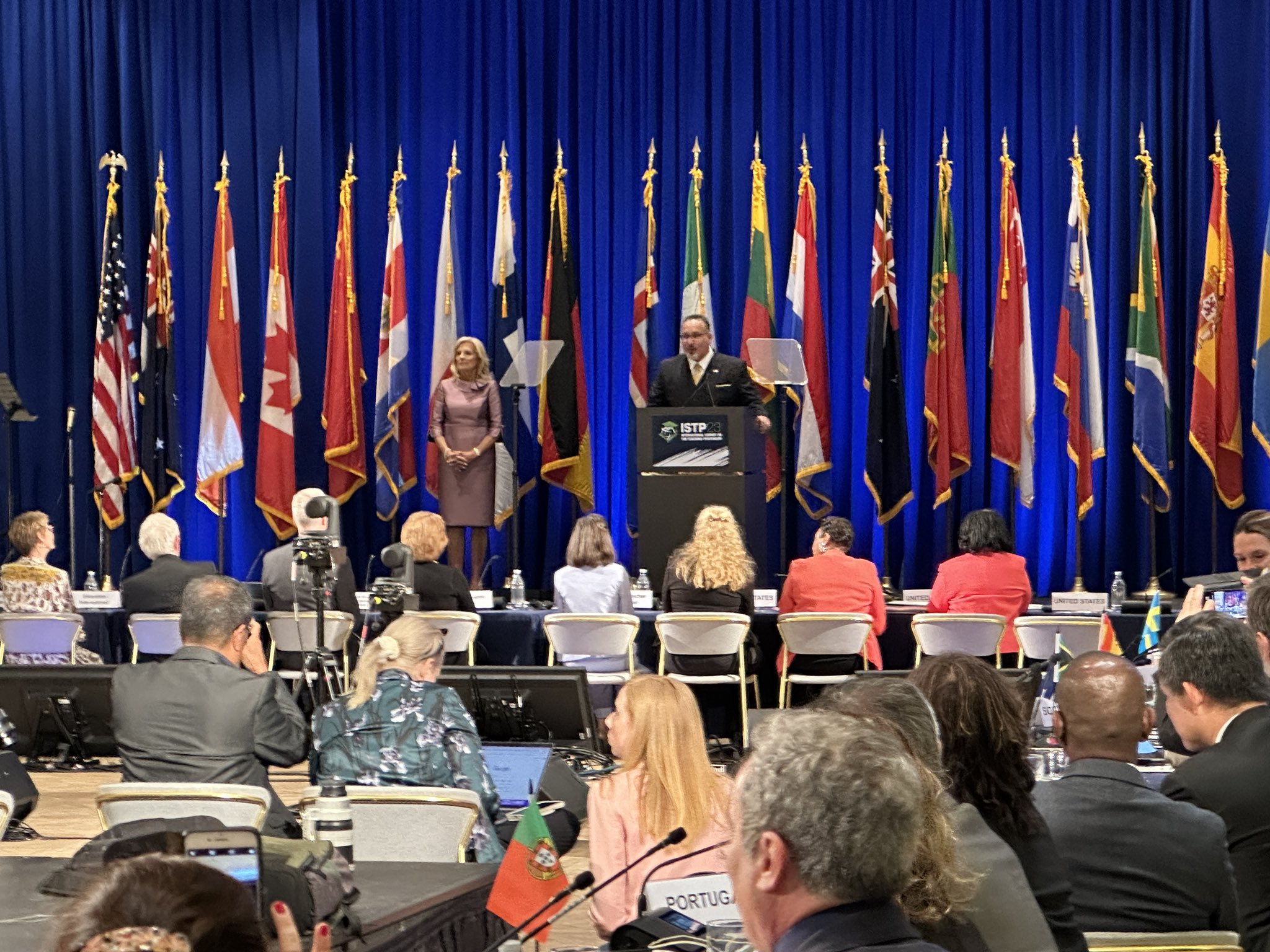 Photo of the First Lady, Education Secretary Miguel Cardona and about 20 other people with international flags in the background.